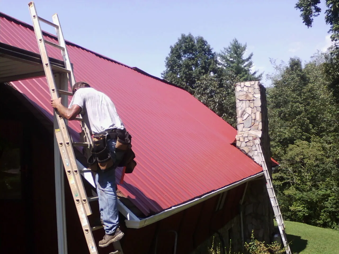 Roofer on a ladder beside a red metal roof and stone chimney — Soffit & Fascia Repair by Roofing Brothers in Darragh